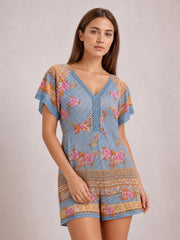 Model poses in a blue floral printed romper with a V-neck, short flutter sleeves, semi-sheer fabric, defined waist, and relaxed shorts silhouette.