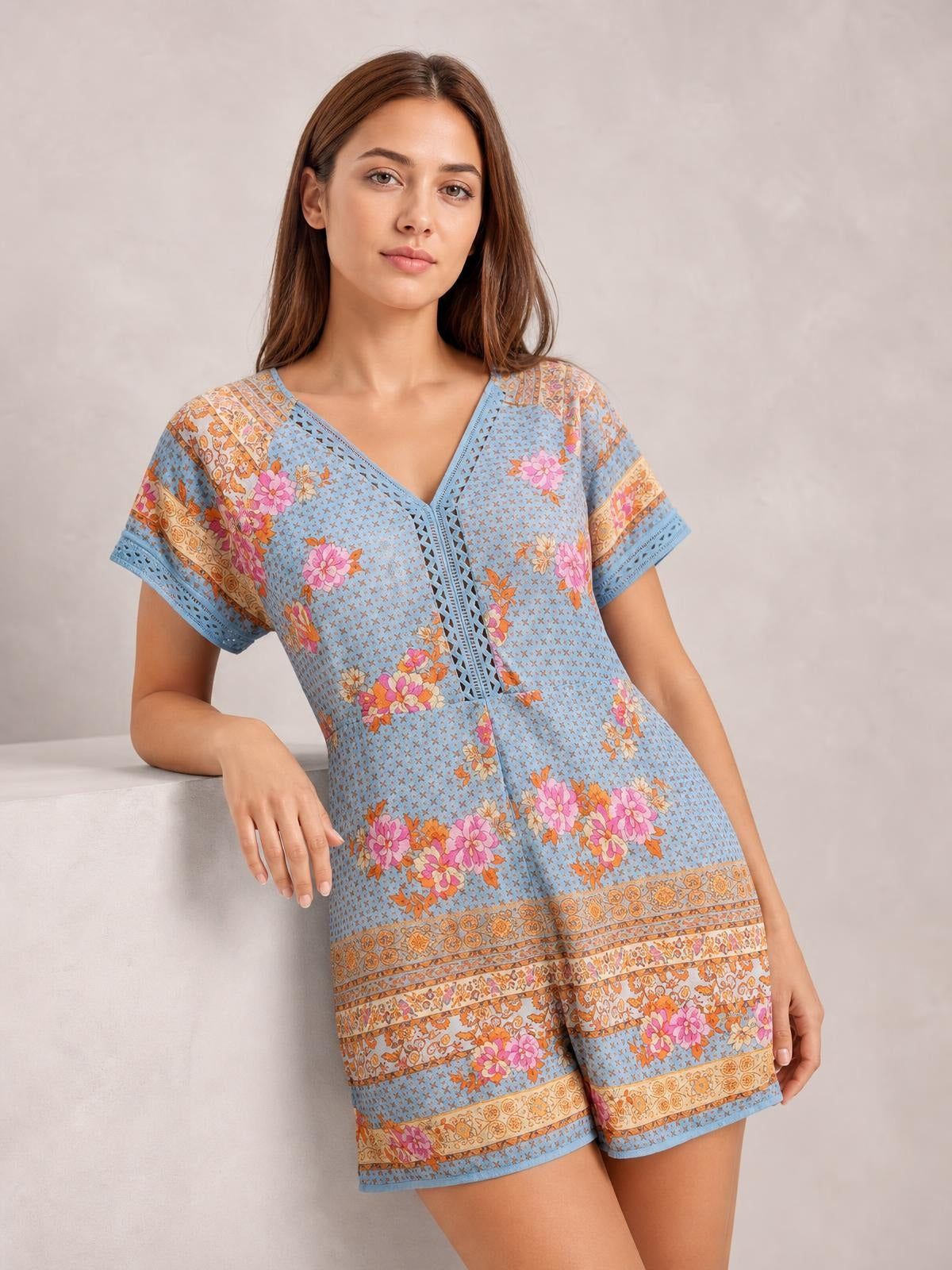 A model leans against a ledge wearing a blue floral printed V-neck short-sleeve romper with a relaxed silhouette, decorative cutout trim, and semi-sheer finish.