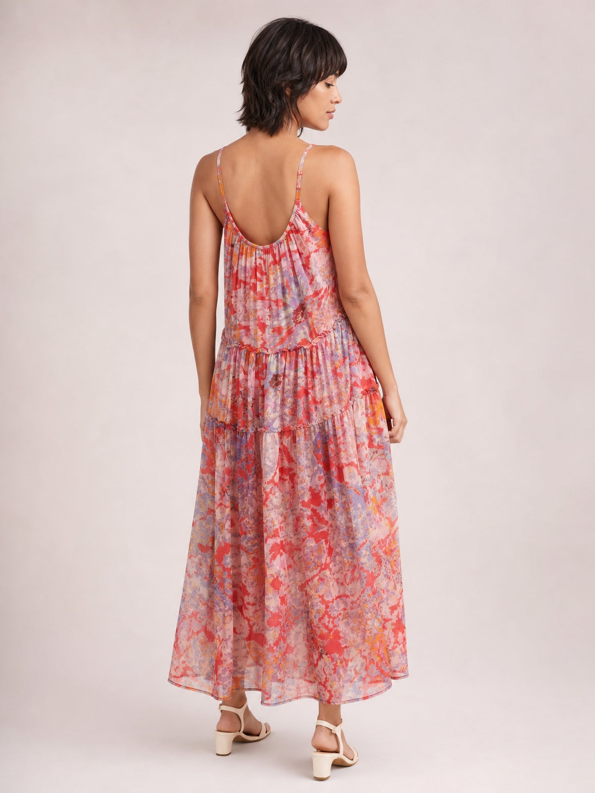 A model turns to show a printed sleeveless dress in coral, pink, and lavender with slender straps, a gathered waist, tiered ankle-length skirt, and flowing silhouette.