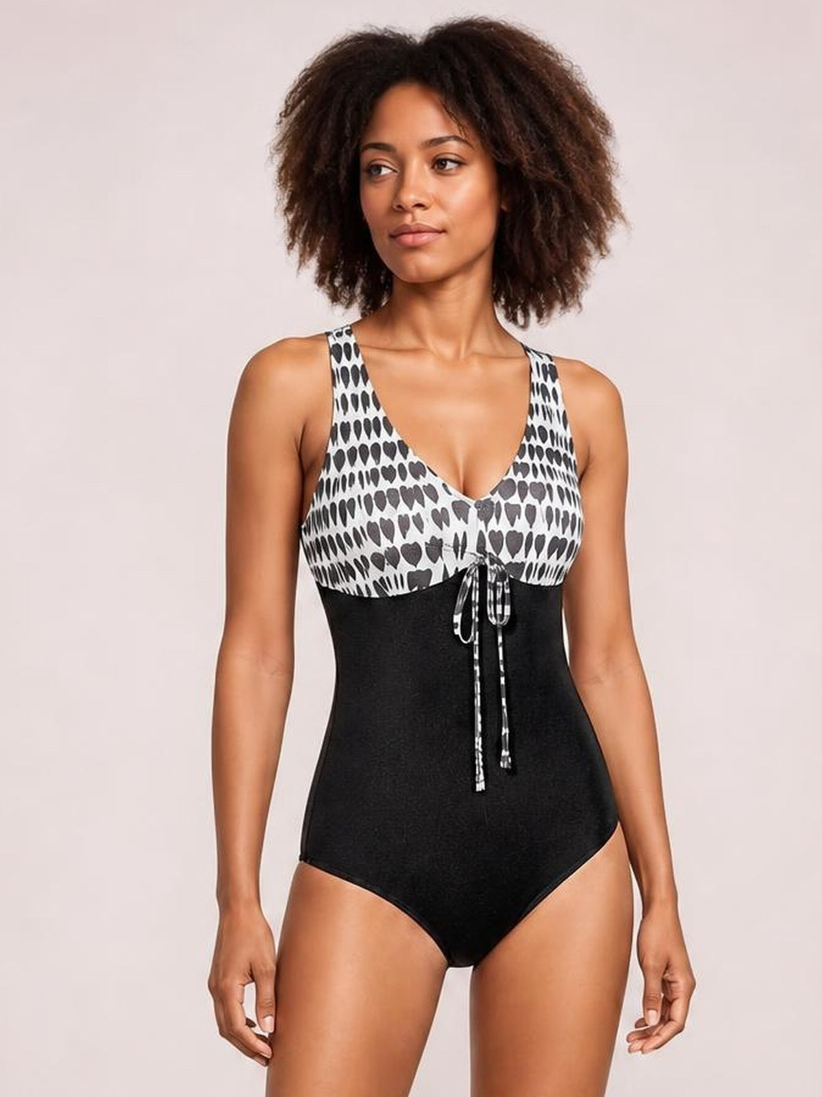 Model stands in a printed black-and-white V-neck sleeveless one-piece swimsuit with wide straps, a black fitted silhouette, and a front tie detail.