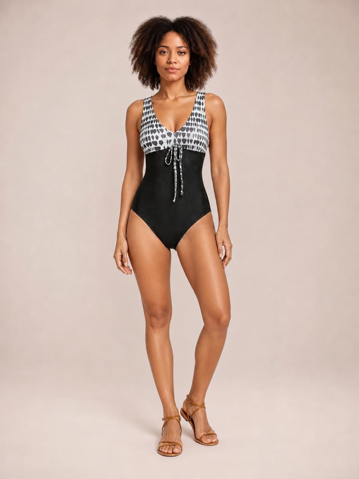 A model stands with one leg crossed forward in a printed V-neck sleeveless one-piece swimsuit with a black fitted silhouette and front tie detail.