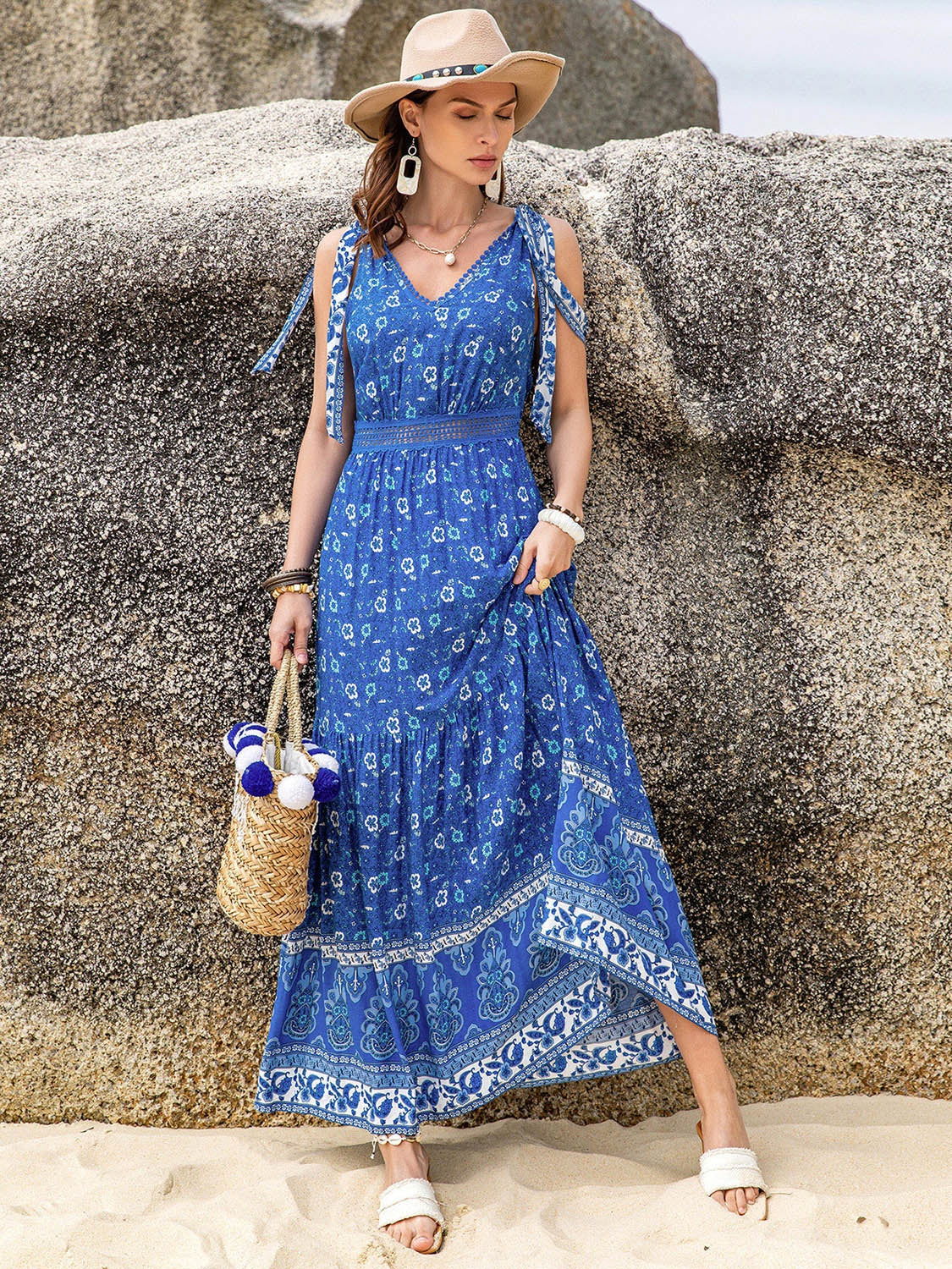 Model poses in a blue printed V-neck tie shoulder dress with a gathered waist, long flowing tiered silhouette, and decorative border print near the hem.