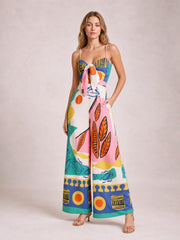 Model poses in a colorful printed cami jumpsuit with tied bust, fitted waist, wide legs, and side pockets.