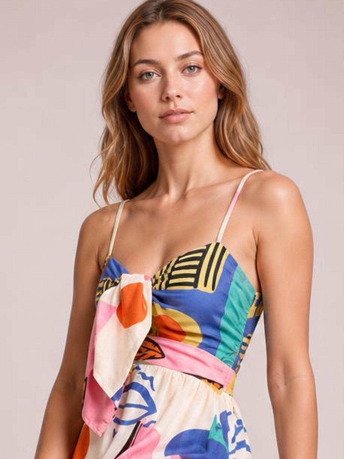 Model wearing a colorful printed wide-leg cami jumpsuit with thin straps, a tied front bodice, fitted waist, and abstract geometric patterns.