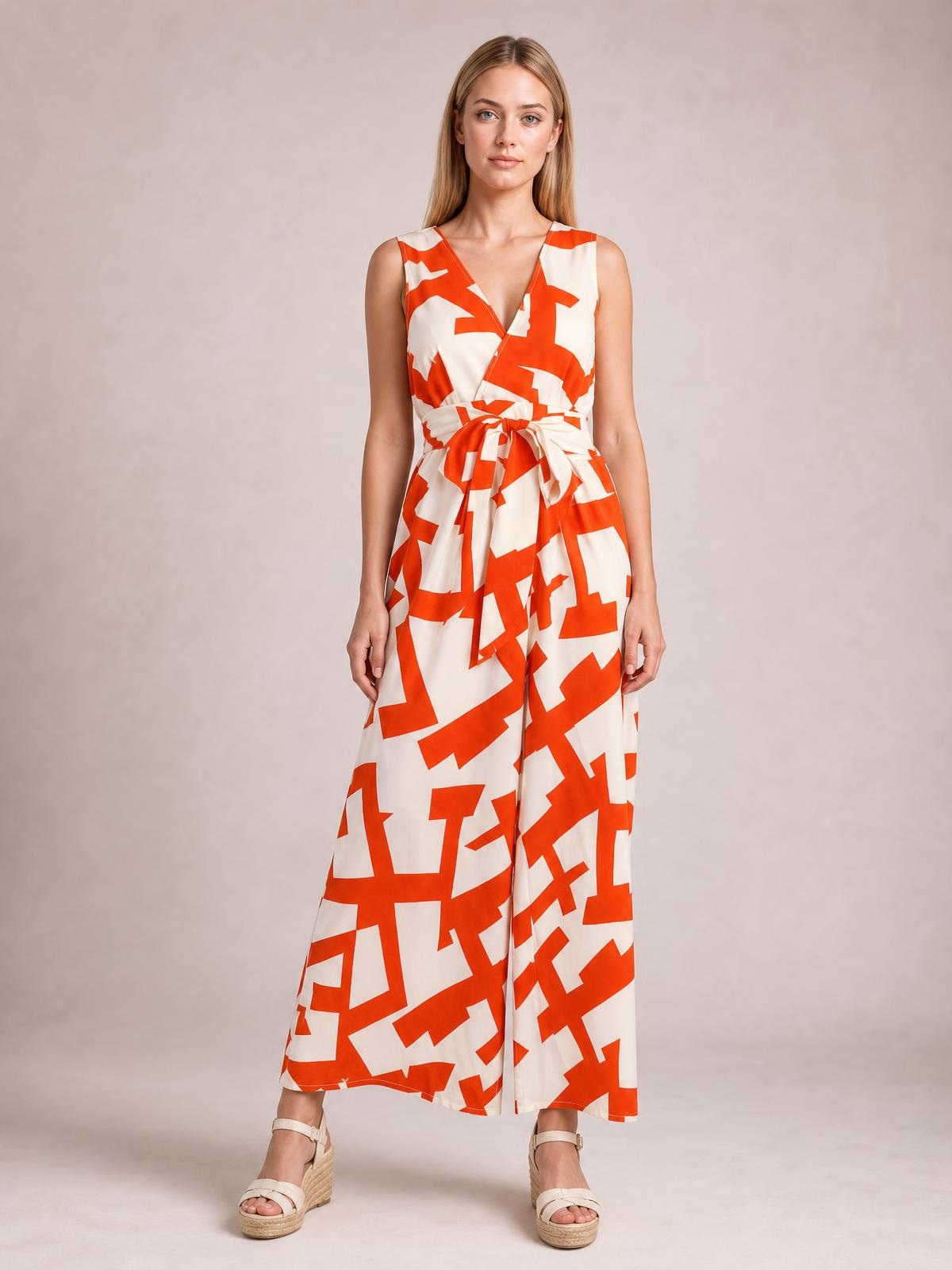 Model stands wearing a printed wide-leg jumpsuit with bold orange and white geometric pattern, sleeveless V-neck bodice, tied waist, and flowing full-length silhouette.