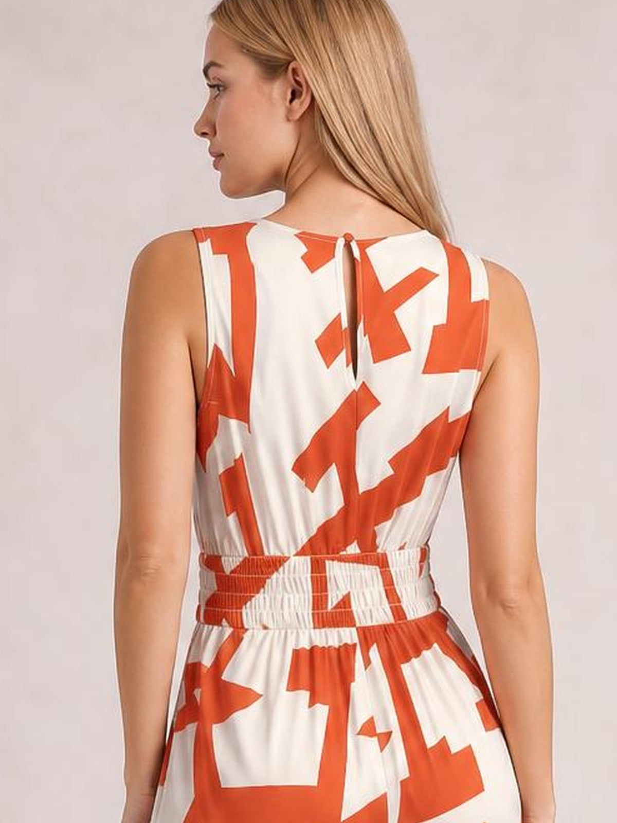 Model turned away to show a white and orange printed wide leg jumpsuit with a sleeveless cut, tied back neckline, and gathered waist.