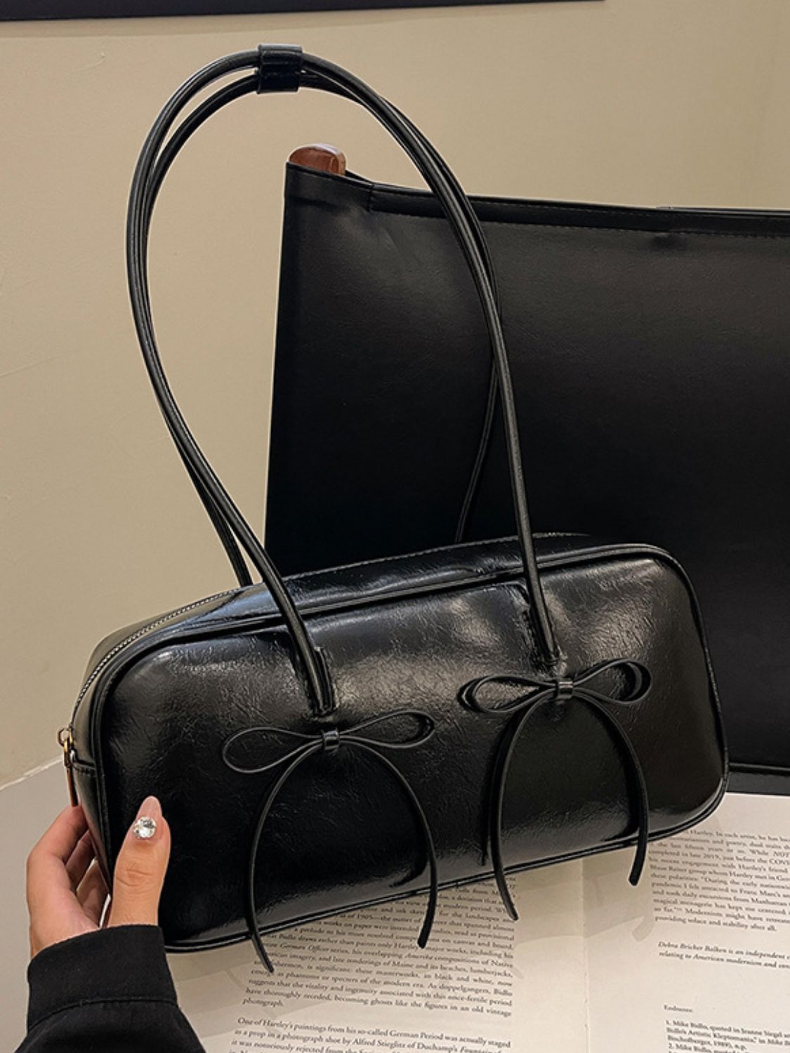 A hand holds a black PU leather shoulder bag with a structured rectangular silhouette, rounded edges, long straps, and two delicate front bow trims.