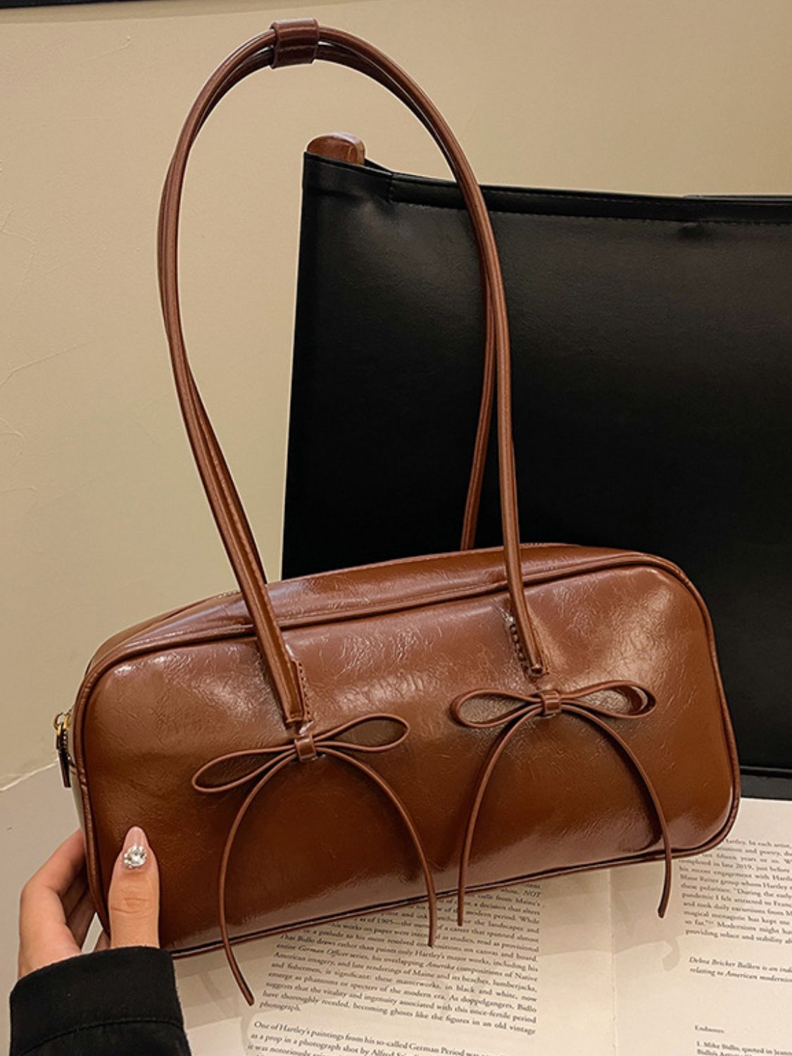 A medium brown PU leather shoulder bag with a structured rectangular silhouette, rounded edges, long straps, polished finish, and twin front bow trims.