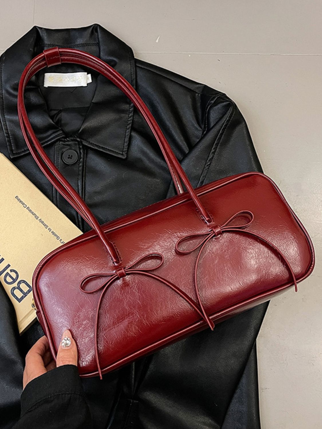 A burgundy PU leather shoulder bag with a structured rounded-rectangle silhouette, long straps, polished edges, and two delicate bow trims across the front.