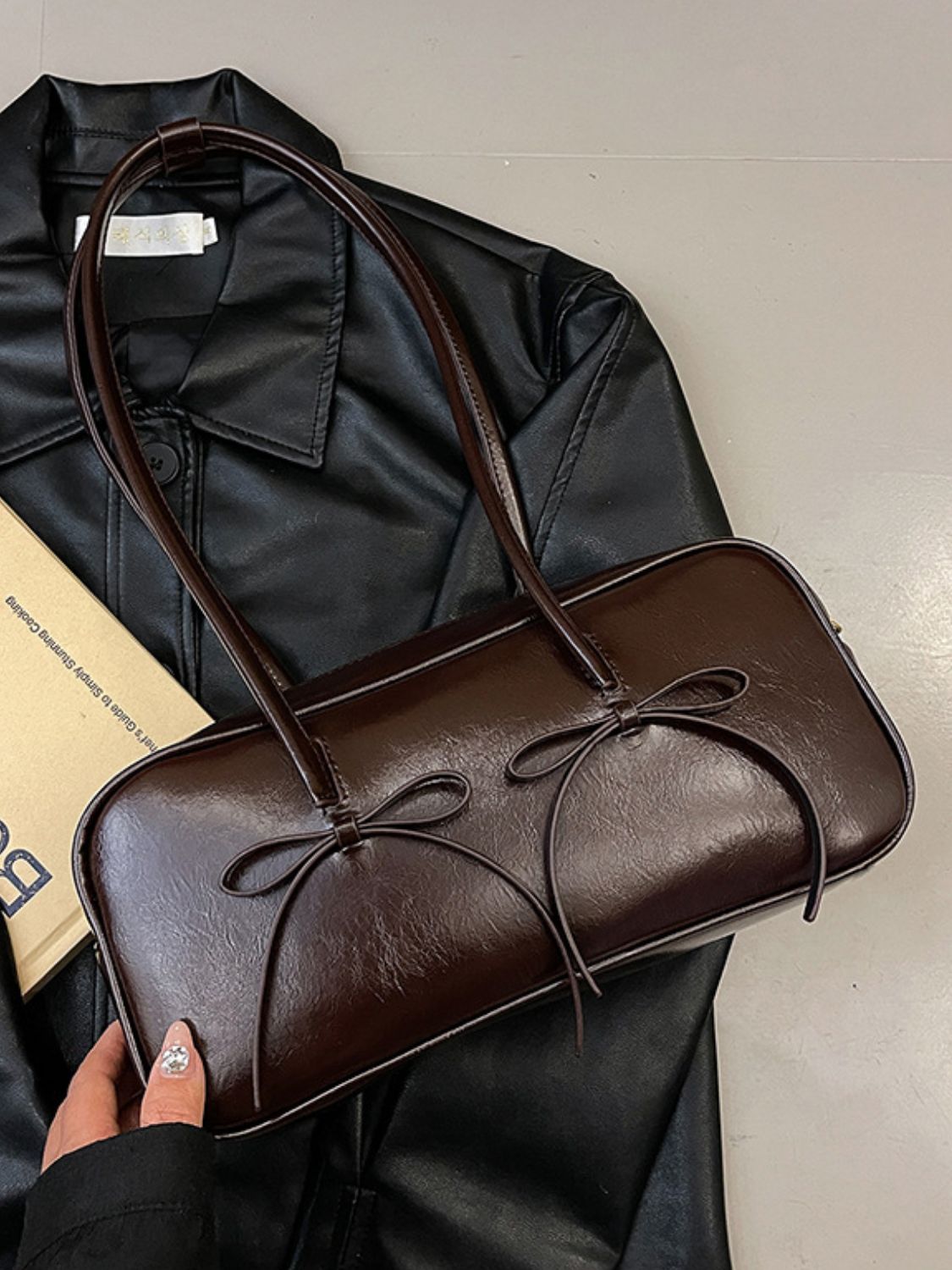 A medium brown PU leather shoulder bag with a structured rectangular silhouette, rounded edges, long straps, and two delicate bow trims across the front.