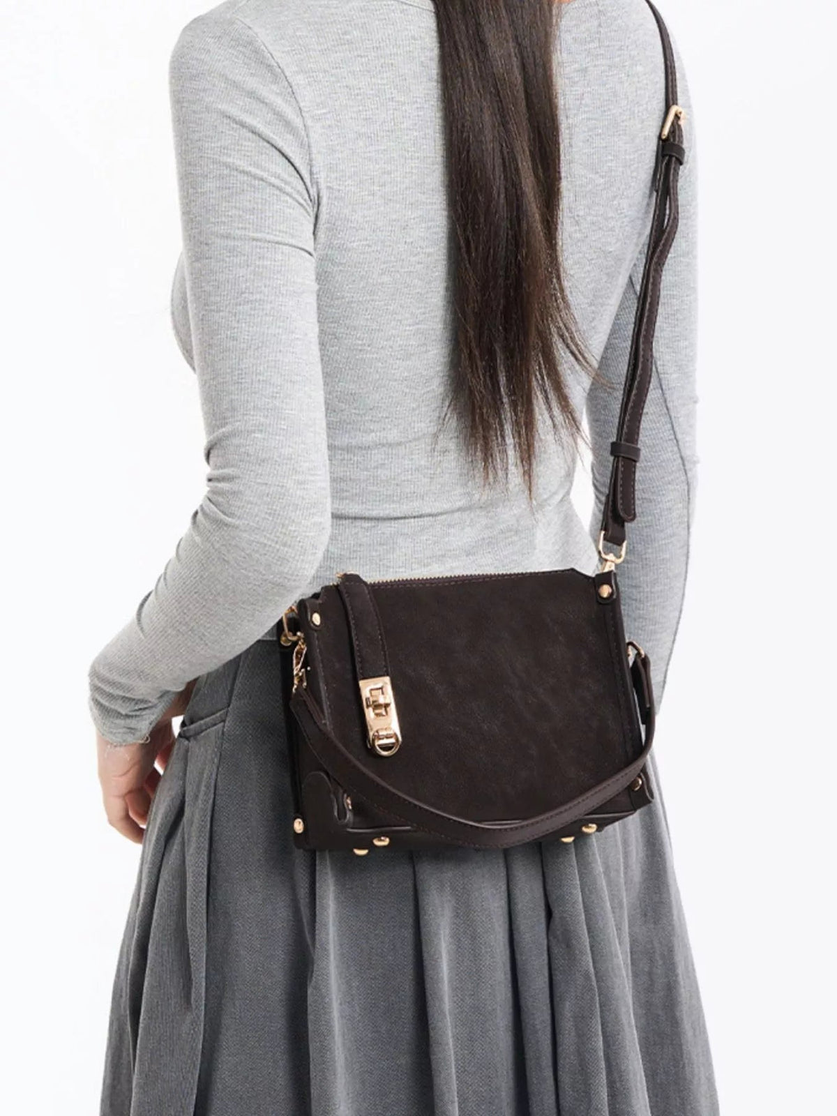 A model wears a fitted gray long-sleeve top and pleated skirt with a small black PU leather crossbody bag, adjustable strap, flap, zipper, and gold-tone hardware.