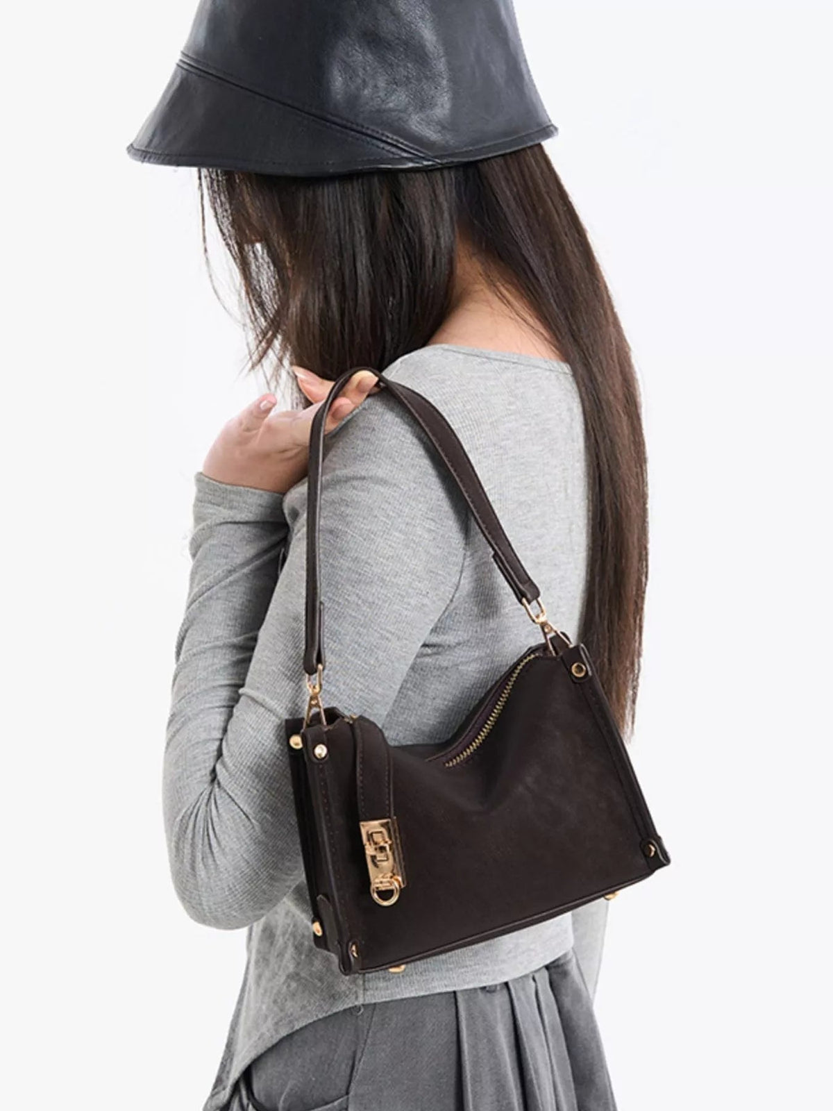 A model carries a small black PU leather crossbody bag with an adjustable strap, structured trapezoid silhouette, top zip, and gold-tone hardware.
