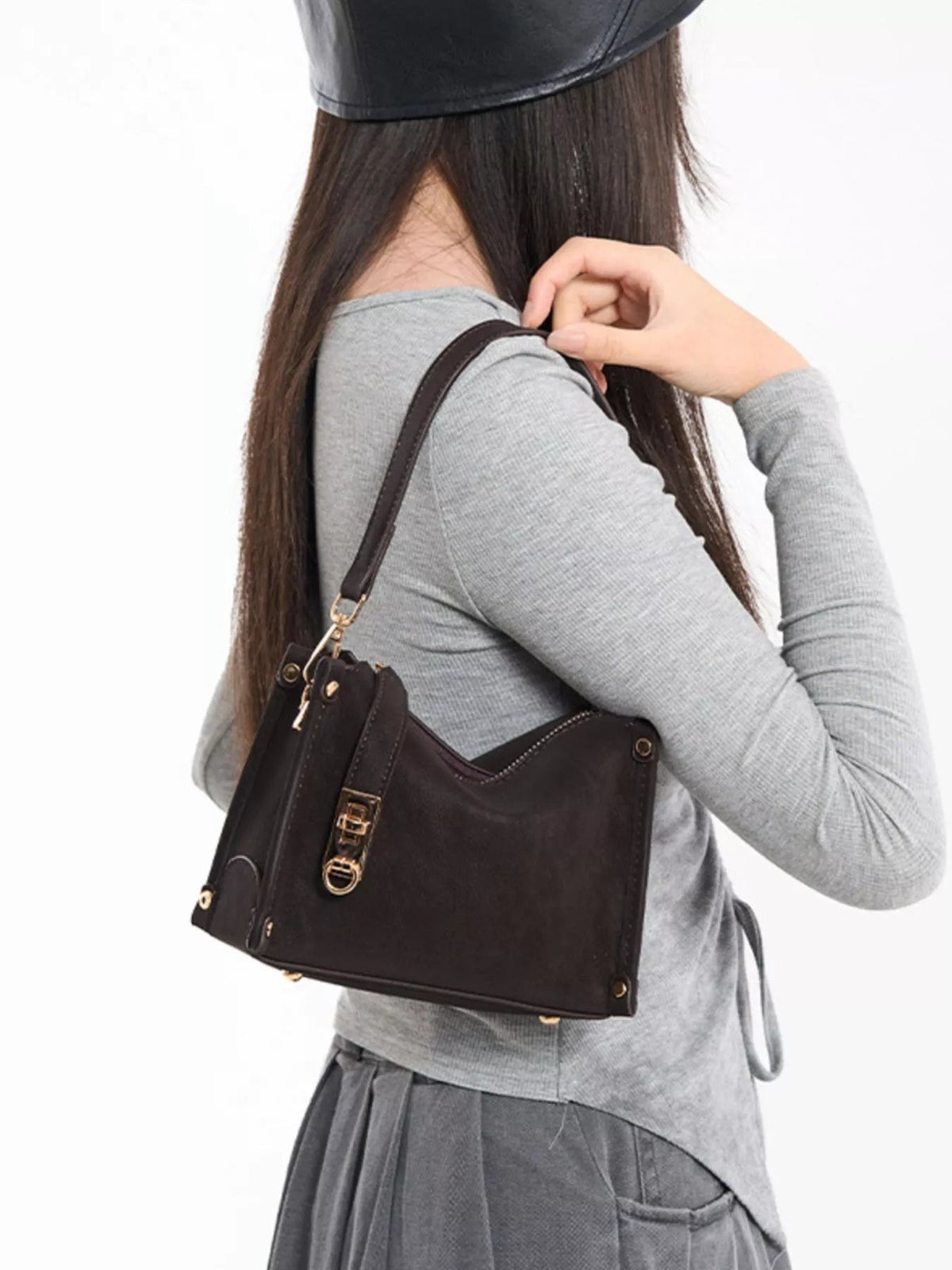A model turned away in a gray long-sleeve top adjusts a small dark brown PU leather bag with an adjustable strap, gold-tone hardware, and zipper detail.
