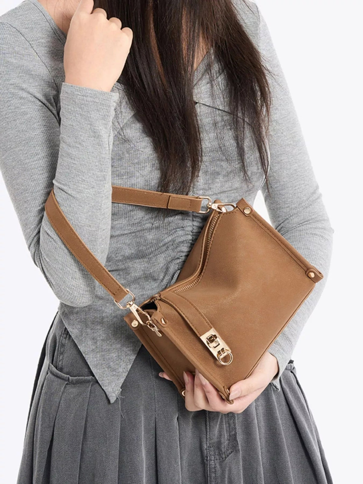A model holds a small brown PU leather crossbody bag with an adjustable strap, structured silhouette, zippered compartments, and gold-tone hardware.