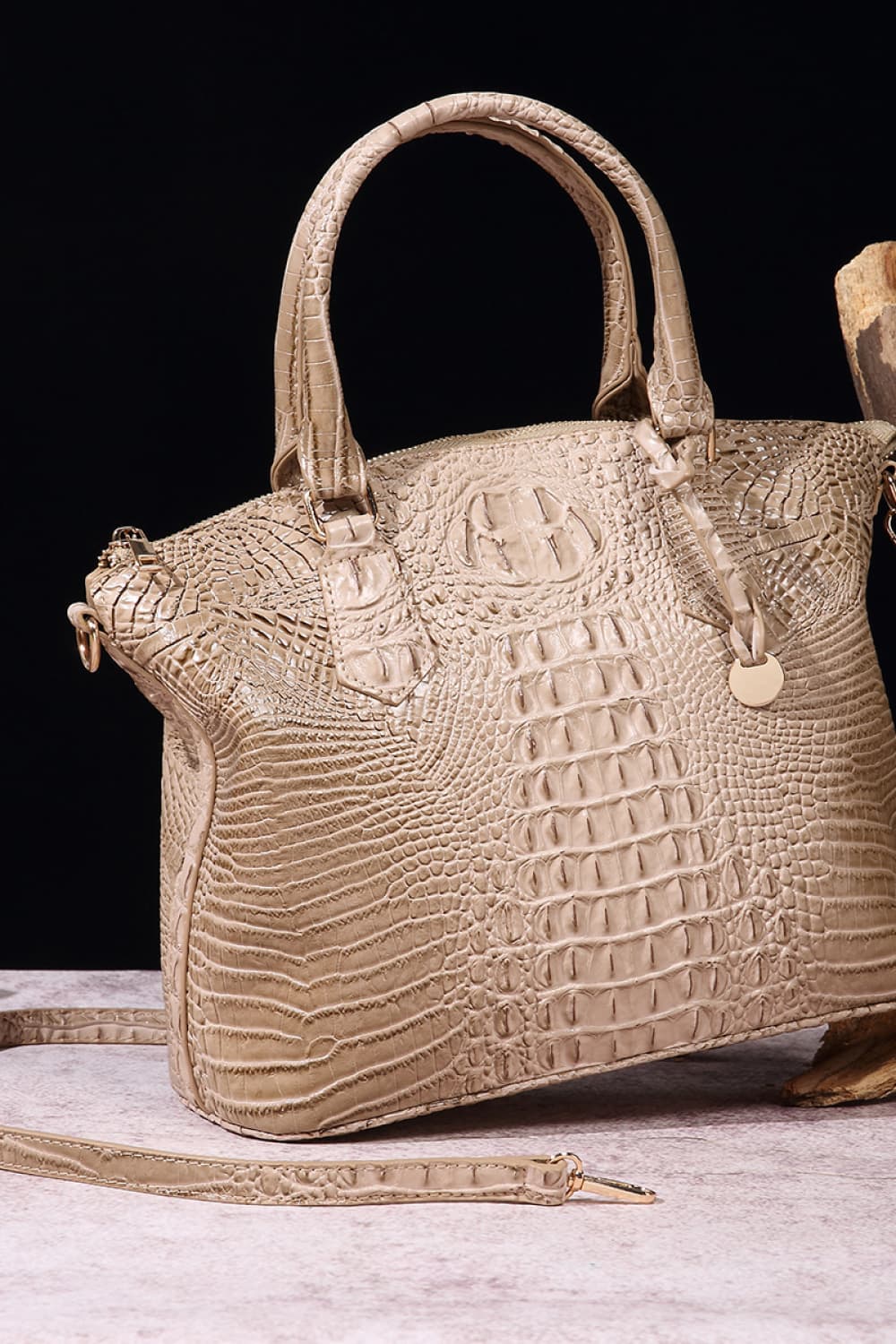 A medium taupe PU leather handbag with a softly tapered silhouette, rounded top handles, crocodile-inspired embossed texture, removable shoulder strap, and hanging charm.