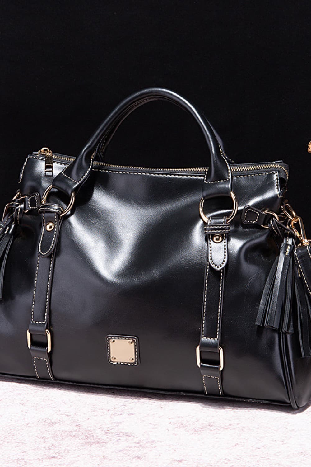 Large black PU leather handbag with rounded top handles, structured spacious silhouette, gleaming hardware, adjustable straps, zip closure, and tassel accents.