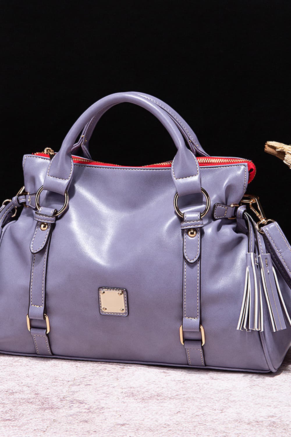 Large lavender PU leather handbag with a softly structured silhouette, rounded top handles, gold-tone hardware, red top zip, adjustable side straps, and tassel accents.