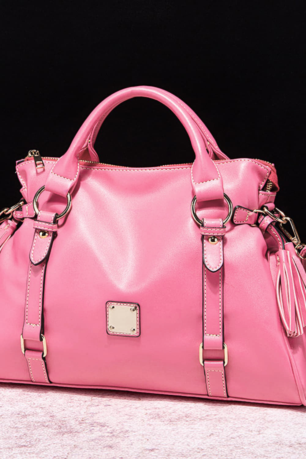 Large pink PU leather handbag with a softly structured silhouette, rounded top handles, adjustable strap details, gleaming hardware, top zip closure, and tassel accents.