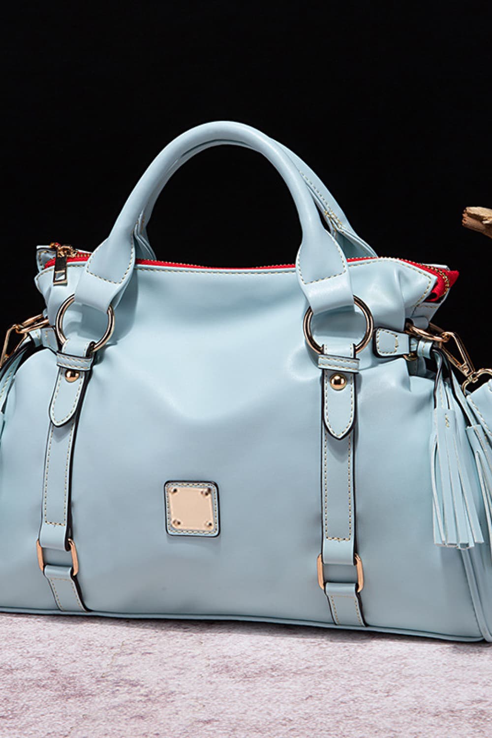 Light blue PU leather handbag with a softly structured silhouette, rounded top handles, gold-tone hardware, front strap details, and a side tassel accent.