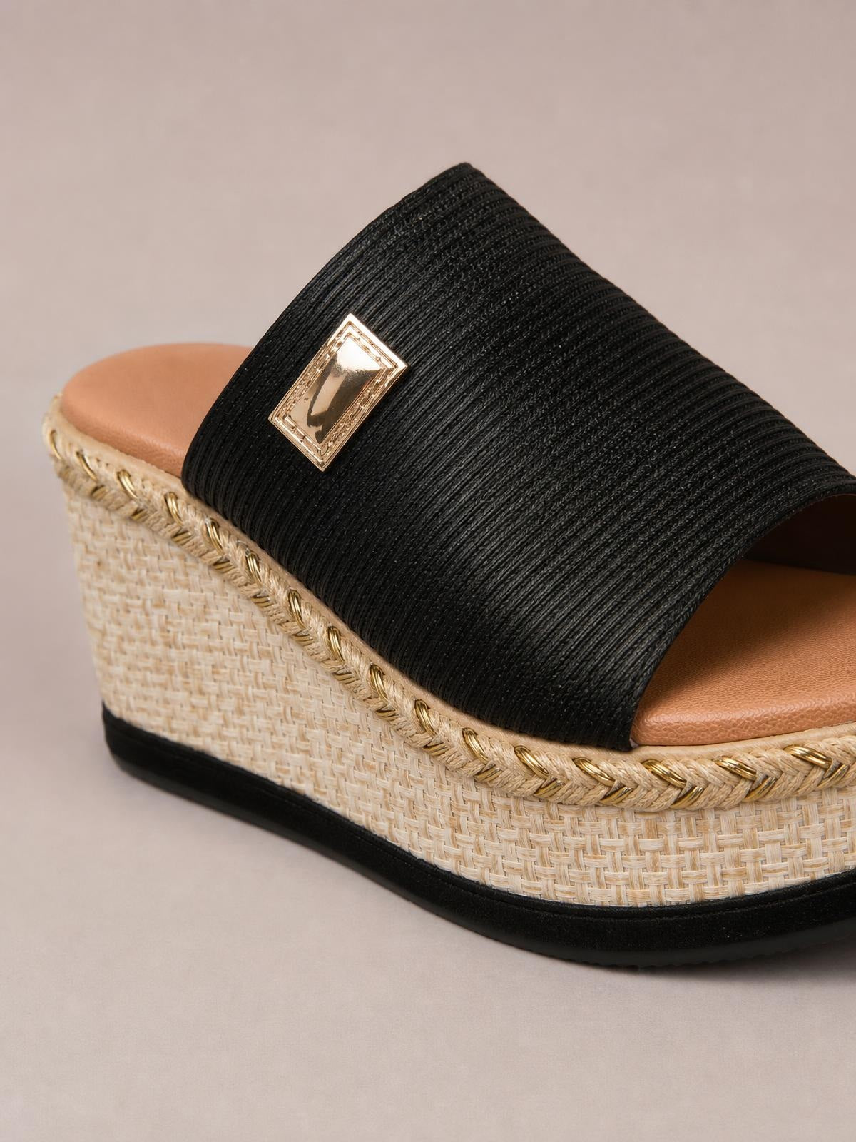 Close-up of a black PU leather open toe sandal with a wide textured strap, gold-tone rectangular ornament, woven wedge platform, and braided trim.