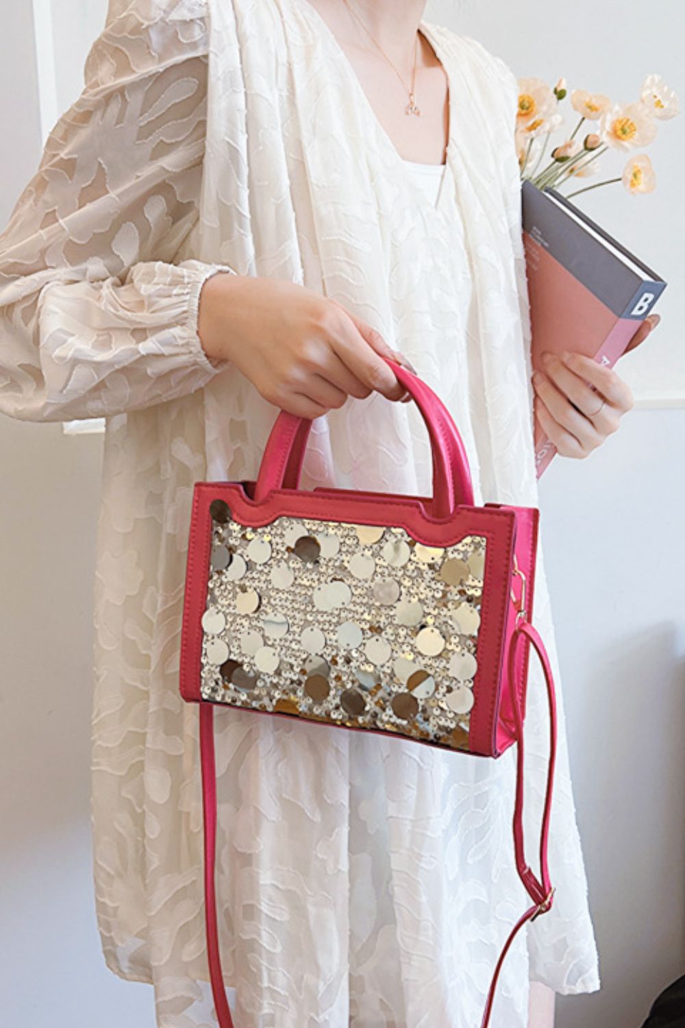 A model holds a small pink PU leather handbag with a structured rectangular silhouette, rounded top handles, a sequin-covered front, and a long shoulder strap.