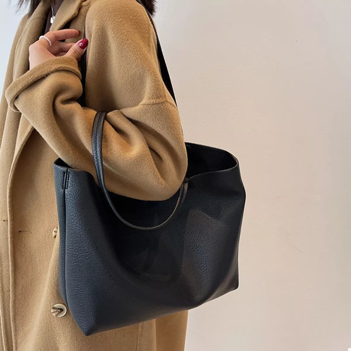 Model carries a large black PU leather tote bag with long shoulder straps, a soft structured silhouette, and an open top.