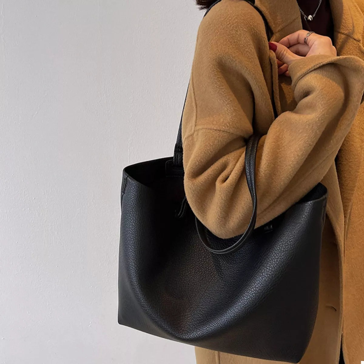 A model carries a large black PU leather tote bag with an open top, pebbled texture, and long shoulder straps over a camel coat.