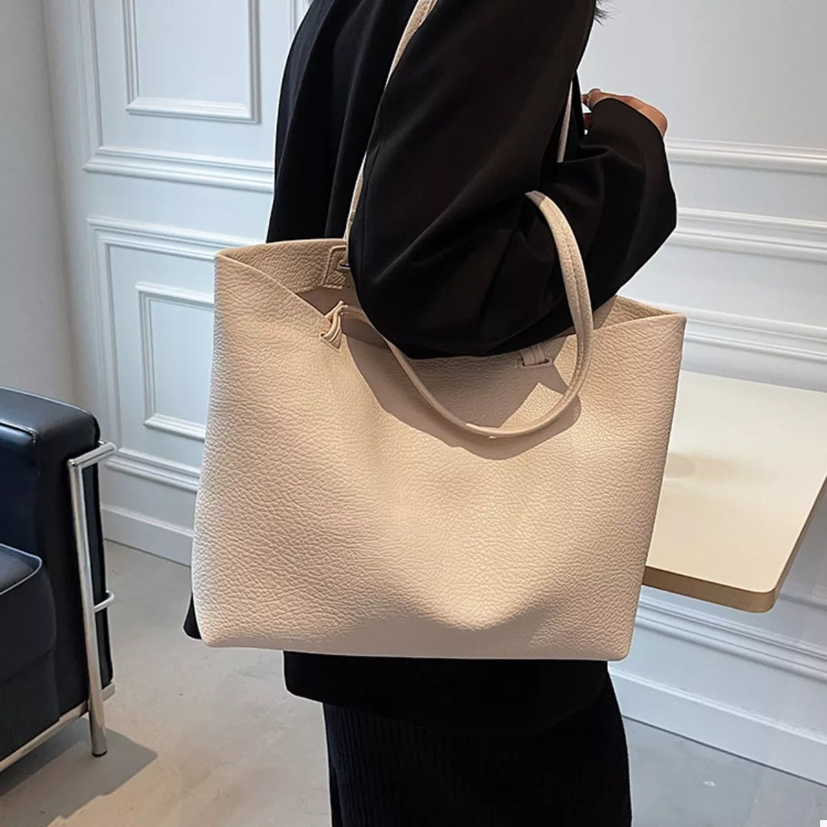 A model in black carries a large beige PU leather tote with a structured rectangular silhouette, pebbled finish, open top, and two rounded handles.