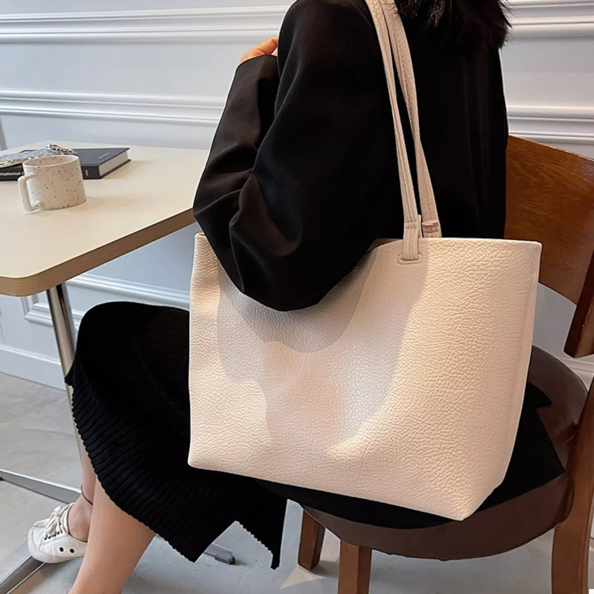 A seated model carries a large cream PU leather tote bag with a pebbled finish, spacious rectangular silhouette, and two long shoulder straps.