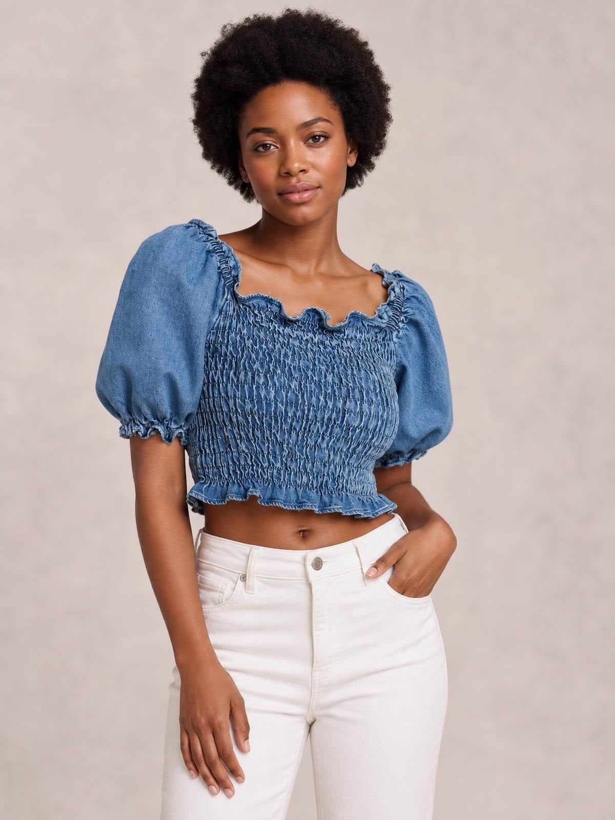 Model poses in a blue cropped denim top with puff sleeves, a smocked bodice, and a ruffled neckline and hem, paired with white pants.