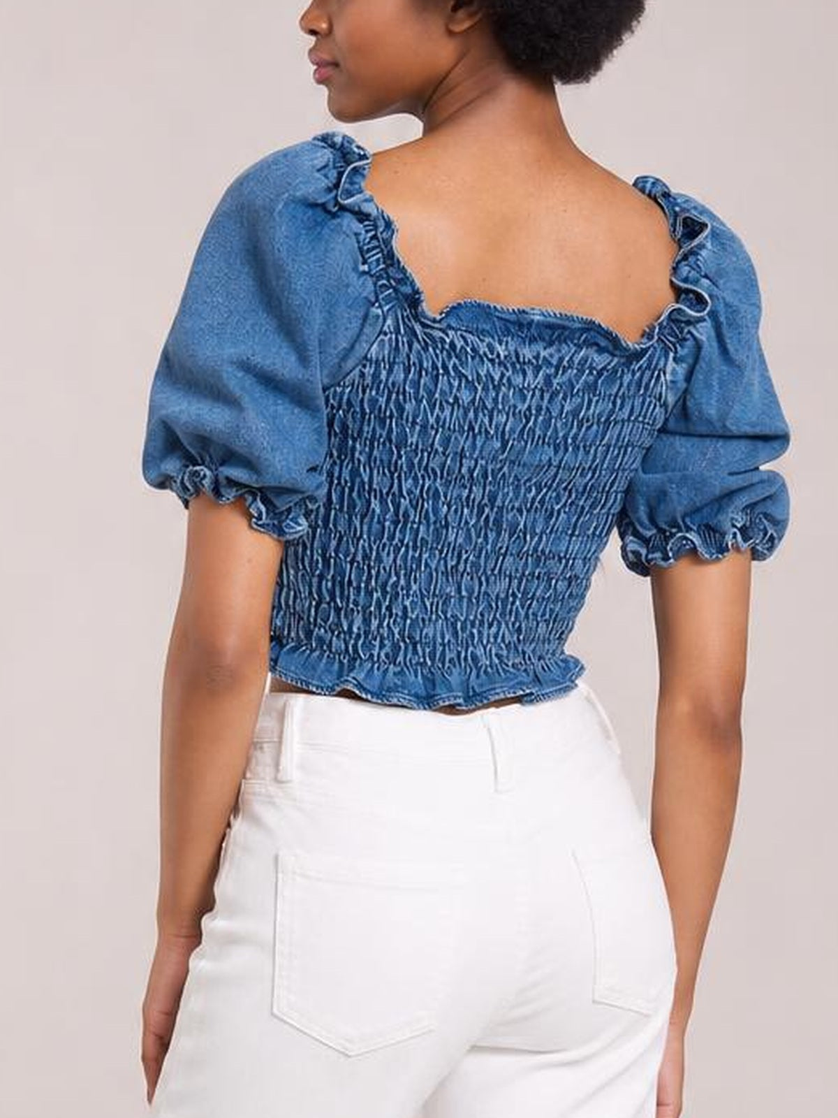 Model shown from the back wearing a blue cropped denim top with a smocked bodice, puff sleeves, gathered trim, and a fitted silhouette.