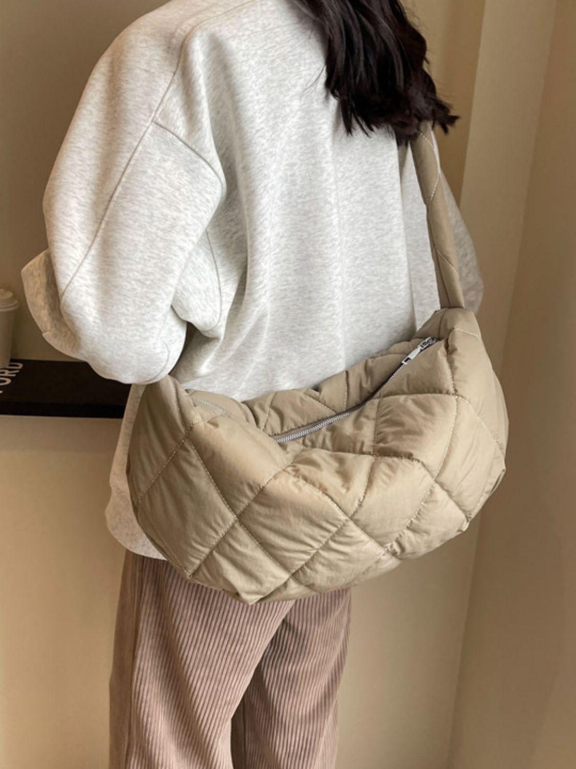 A model wears a large beige quilted crossbody bag with a softly rounded crescent silhouette, plush volume, long strap, and visible zip-top closure.