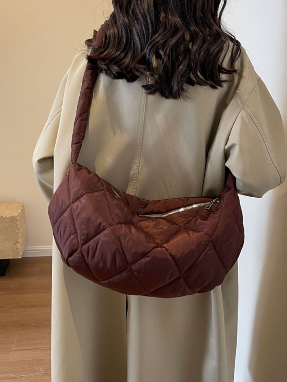 A model wears a large burgundy quilted crossbody bag with a softly rounded crescent silhouette, plush volume, long strap, and zip-top closure.