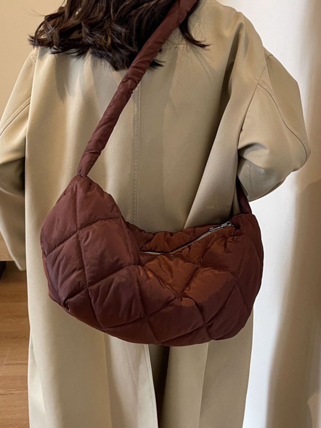 A model wears a large burgundy quilted crossbody bag with a softly structured crescent silhouette, plush volume, long strap, and zip-top closure.