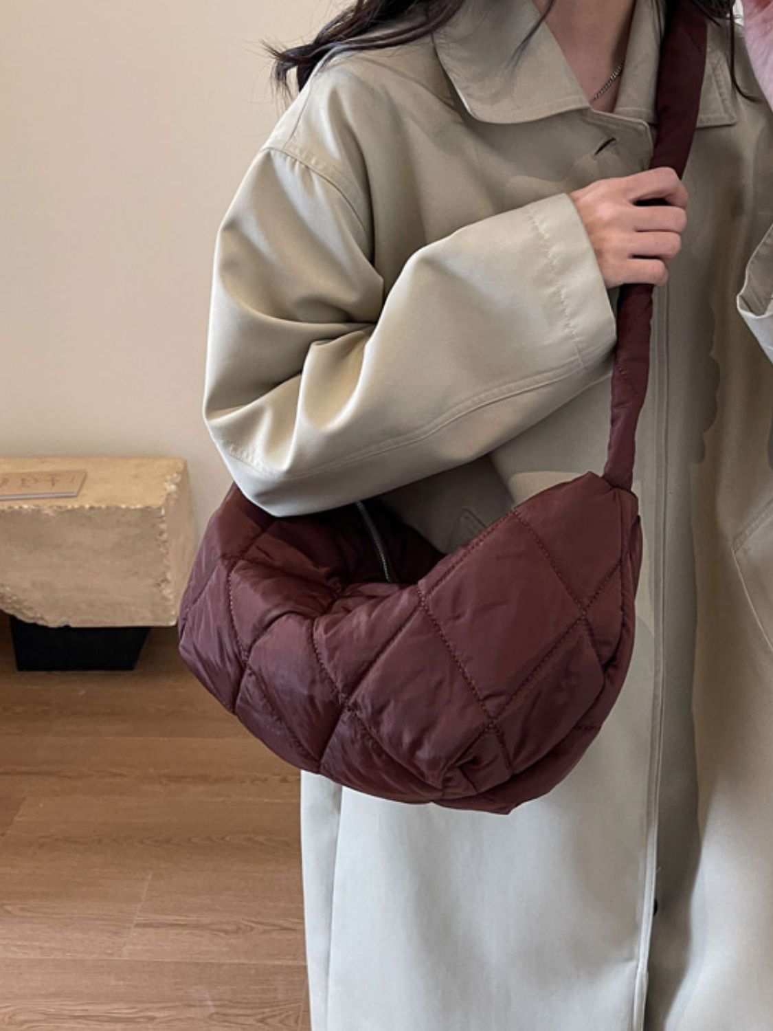 A model holds a large burgundy quilted crossbody bag with a plush crescent silhouette and long strap worn diagonally across the torso.