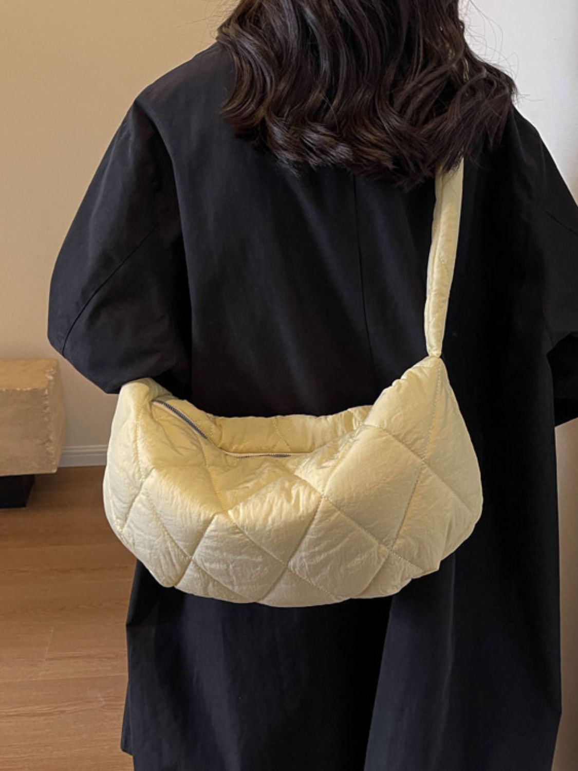 Model wearing a large cream quilted crossbody bag with a softly structured crescent silhouette, plush volume, long strap, and zip-top opening.