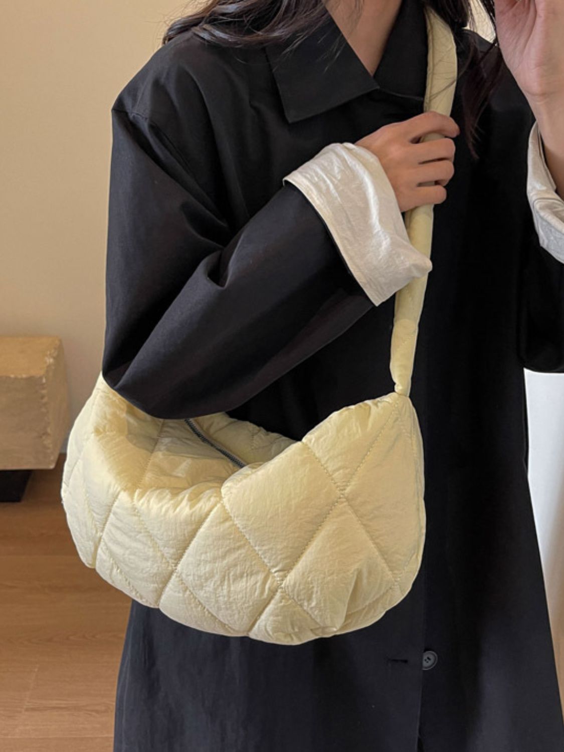 Model wearing a black collared coat carries a large cream quilted crossbody bag with a softly structured crescent silhouette and long strap.