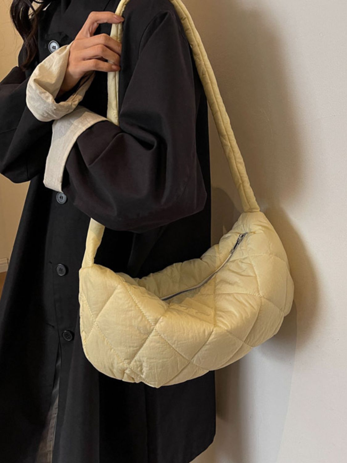 Model wears a large cream quilted crossbody bag with a softly rounded crescent silhouette, plush volume, long strap, and visible zip-top closure.