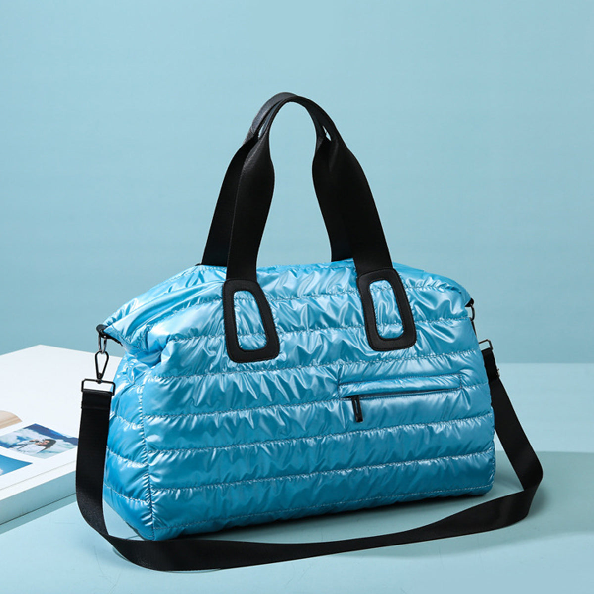 Oversize blue quilted nylon travel bag with a softly padded silhouette, black top handles, detachable shoulder strap, and front zip pocket.