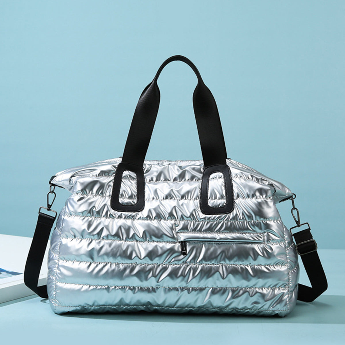 An oversize silver quilted nylon travel bag with a softly padded silhouette, black top handles, detachable shoulder strap, and front zip pocket.