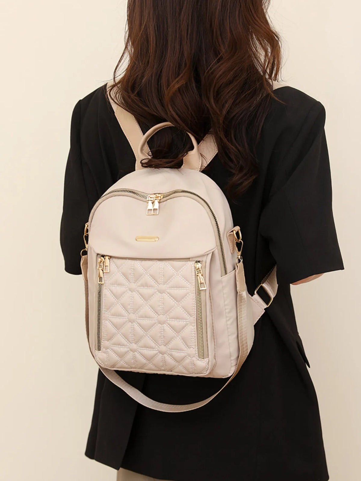 A model wears a medium light beige quilted Oxford cloth backpack with adjustable shoulder straps, gold-tone zippers, and a diamond-stitched front pocket.