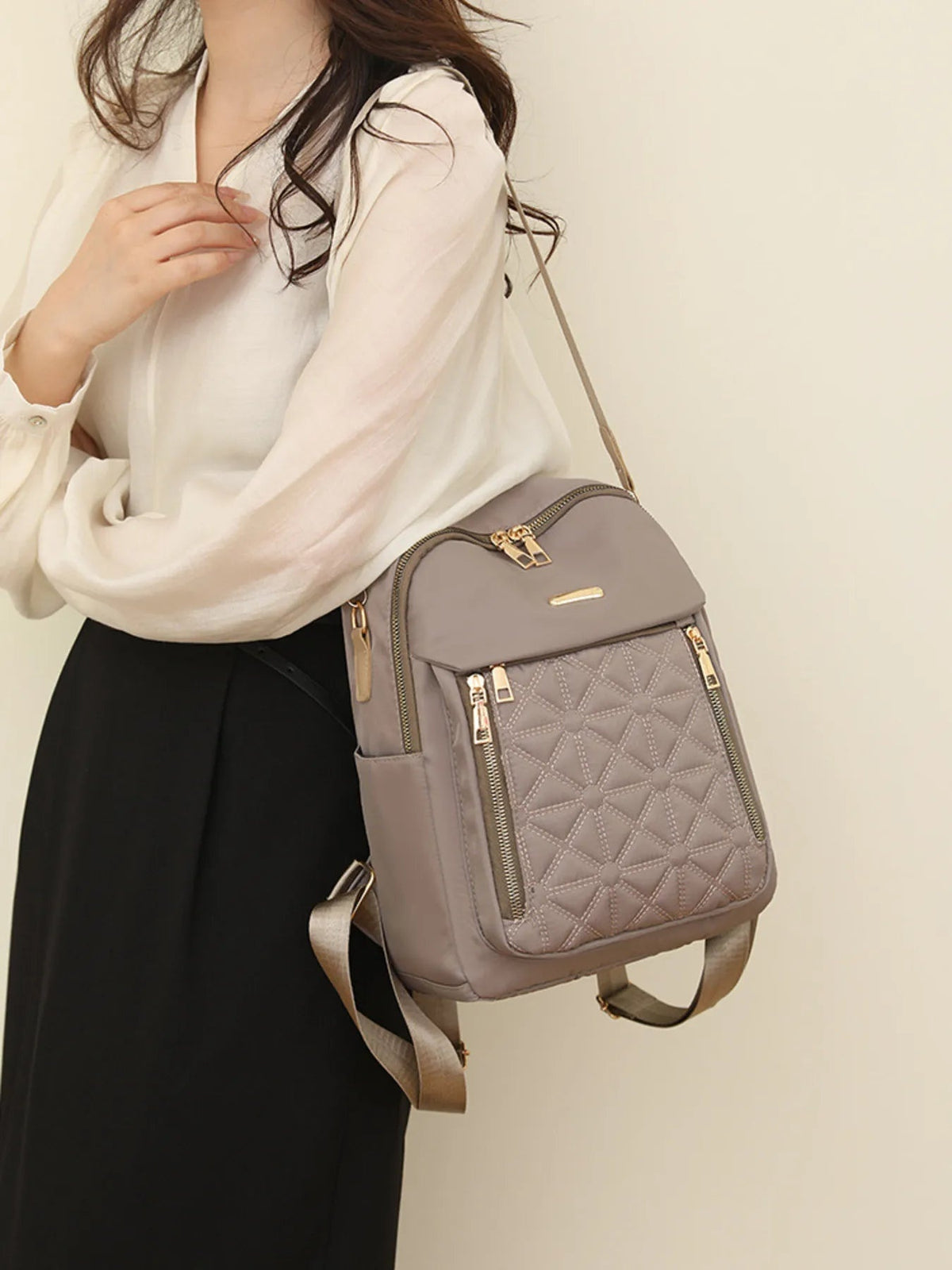 A model wears a medium taupe quilted Oxford cloth backpack with a structured silhouette, front zip pockets, gold-tone hardware, and adjustable shoulder straps.