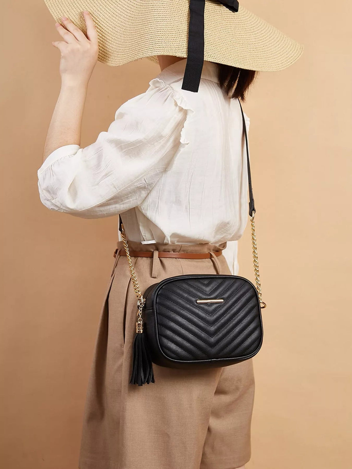 A model wears a black mini PU leather crossbody bag with chevron quilting, rounded rectangular shape, gold chain strap, and a long tassel at her hip.