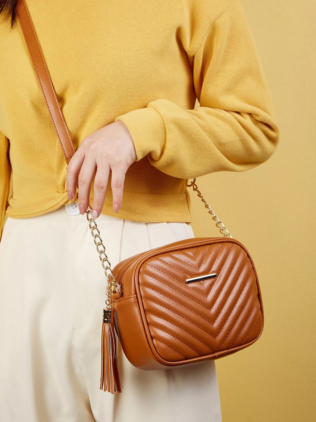 Model wearing a mini brown PU leather crossbody bag with chevron quilting, a chain strap, structured rounded silhouette, and long tassel detail.