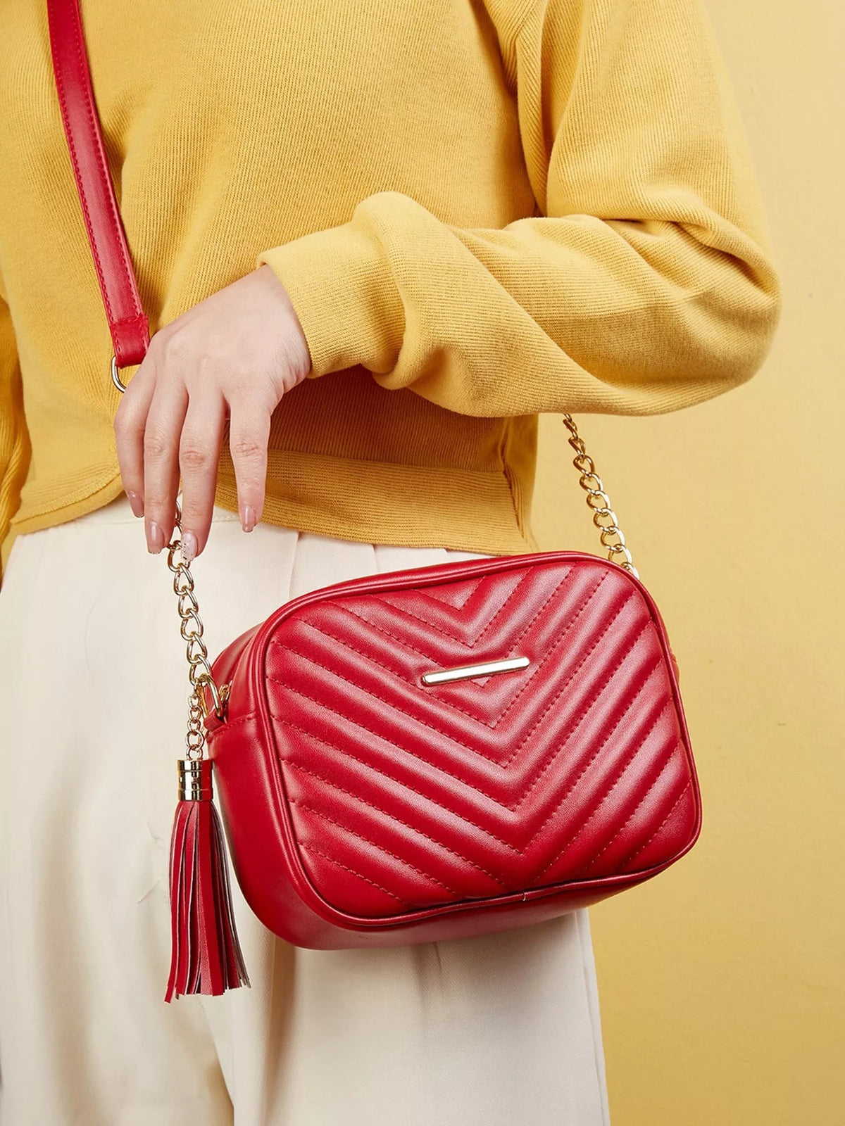 A model wears a red quilted PU leather mini crossbody bag with chevron stitching, a rounded rectangular silhouette, gold chain strap, and tassel.