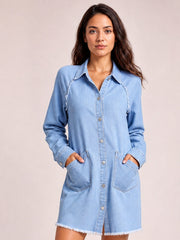 Model poses with hands in the pockets of a light blue raw hem button-up denim dress featuring a collared neckline, long sleeves, relaxed silhouette, and front pockets.