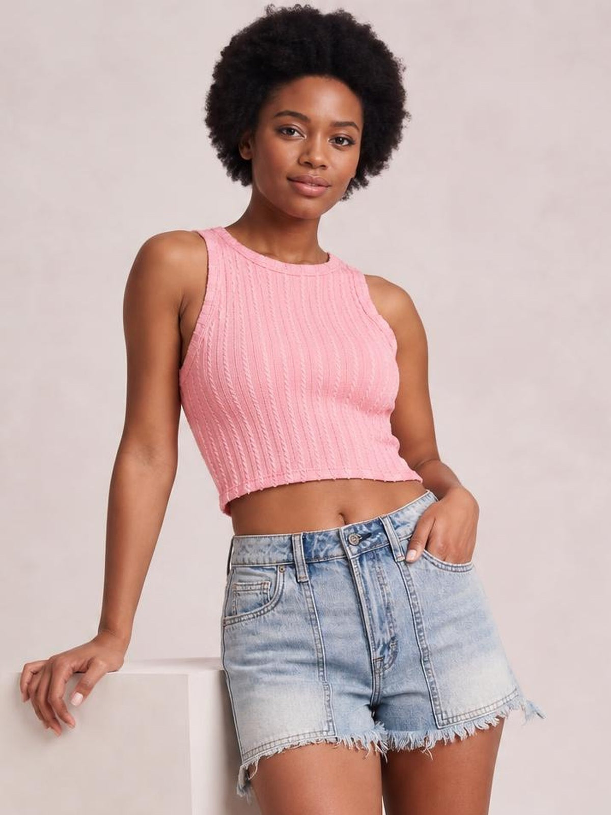 A woman wearing a pink ribbed knit sleeveless crop top and light-wash denim shorts, posing against a neutral beige studio background.