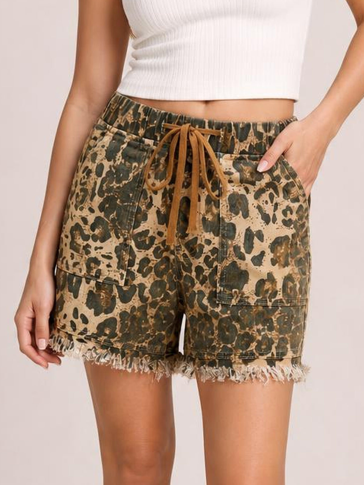 A model wears leopard-print denim shorts with a relaxed silhouette, drawstring waist, front pockets, and frayed raw hems, standing with one hand tucked into a pocket.