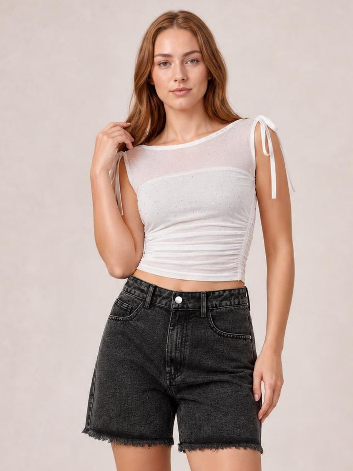 A woman modeling a white ribbed crop top with sheer mesh shoulders and tie details, paired with black denim shorts, against a neutral beige background.