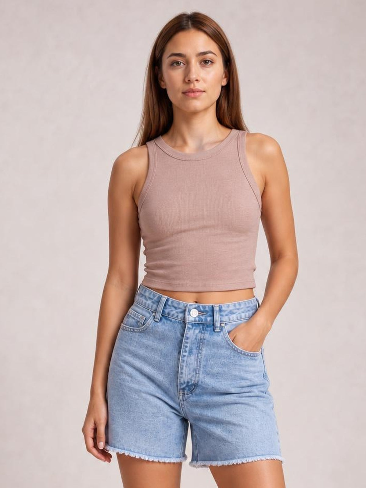A woman modeling a sleeveless ribbed crop top in a dusty mauve-pink color paired with light-wash denim shorts, set against a neutral textured beige background.